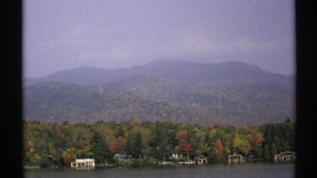 1971 New York home-movie view of lakefront cottages among fall trees, with a hazy wooded mountain range in the background.