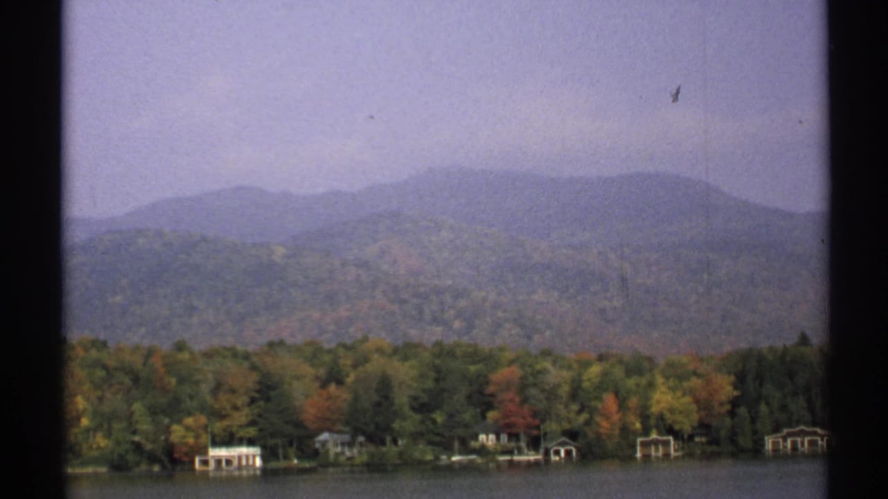 1971 New York 8mm frame showing a wooded lakeshore with autumn color, small waterside structures, distant hills, and soft haze.