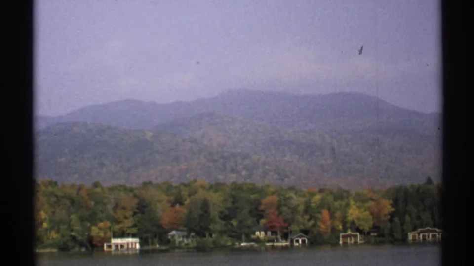 1971 New York 8mm frame showing a wooded lakeshore with autumn color, small waterside structures, distant hills, and soft haze.