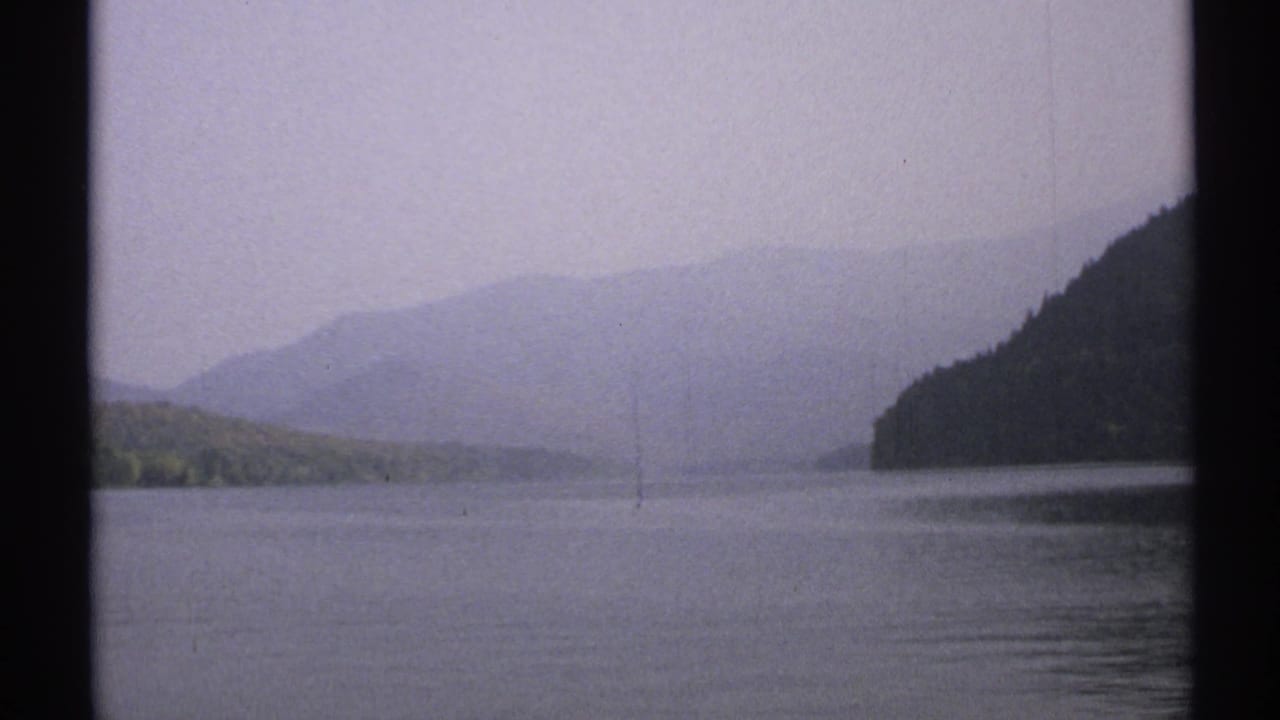 1971 New York 8mm frame looking across calm lake water toward wooded slopes and layered hills under an overcast, hazy sky.