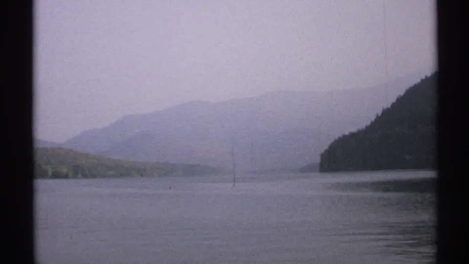 1971 New York 8mm frame looking across calm lake water toward wooded slopes and layered hills under an overcast, hazy sky.