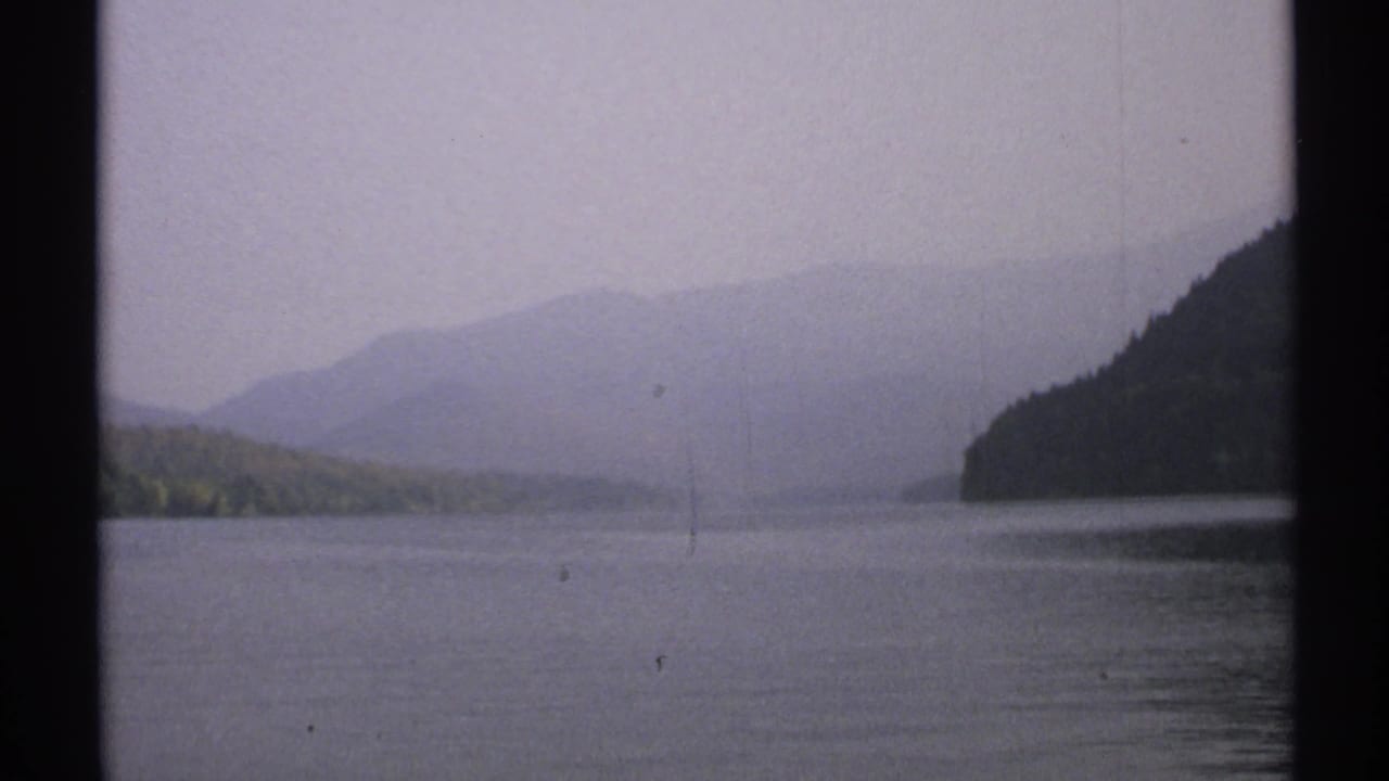 1971 New York 8mm frame of calm lake water, a low distant shoreline, hazy hills, and a dark wooded slope along the right edge.