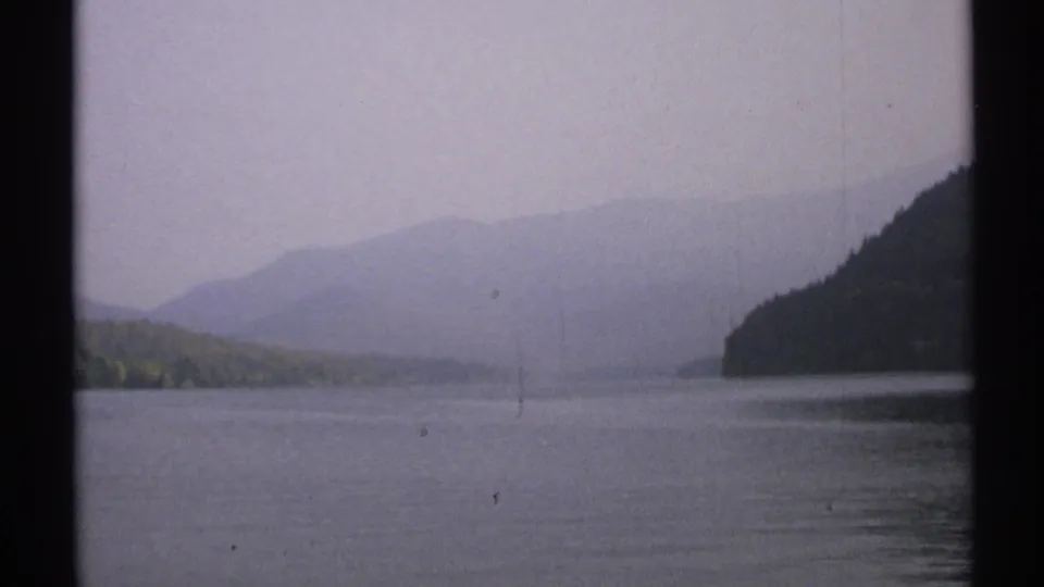1971 New York 8mm frame of calm lake water, a low distant shoreline, hazy hills, and a dark wooded slope along the right edge.