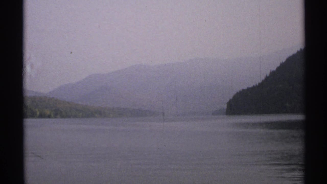 1971 New York 8mm frame showing a tranquil lake, low wooded shoreline, hazy hills, muted sky, and visible film-frame borders.