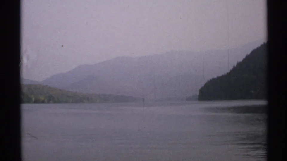 1971 New York 8mm frame showing a tranquil lake, low wooded shoreline, hazy hills, muted sky, and visible film-frame borders.