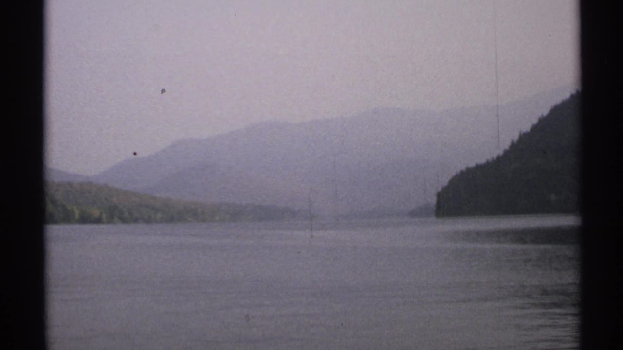 1971 New York 8mm view across a misty lake toward wooded hills and faint distant mountains, seen through dark vertical framing.