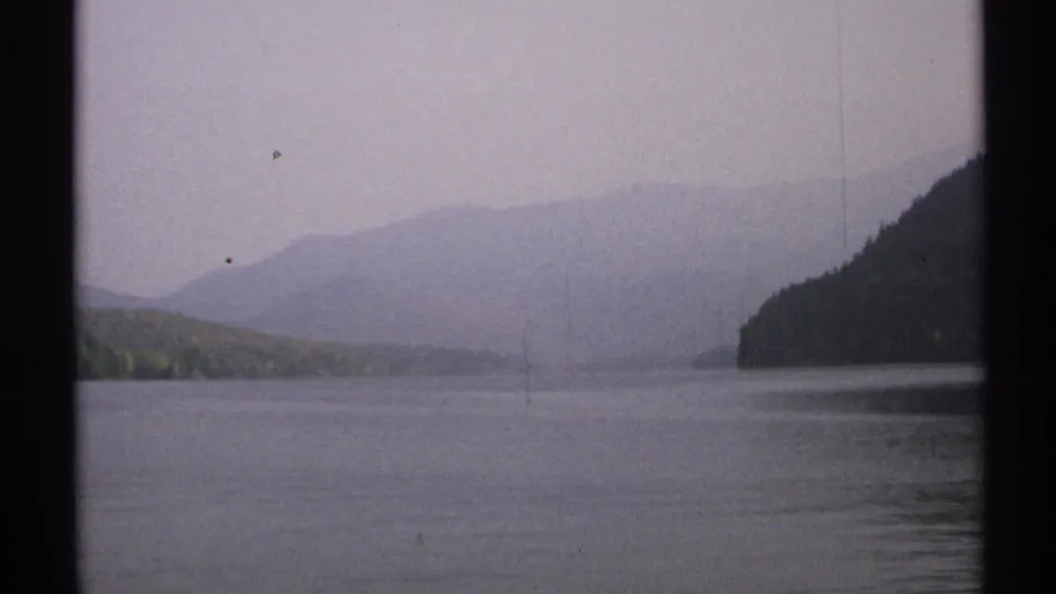 1971 New York 8mm view across a misty lake toward wooded hills and faint distant mountains, seen through dark vertical framing.