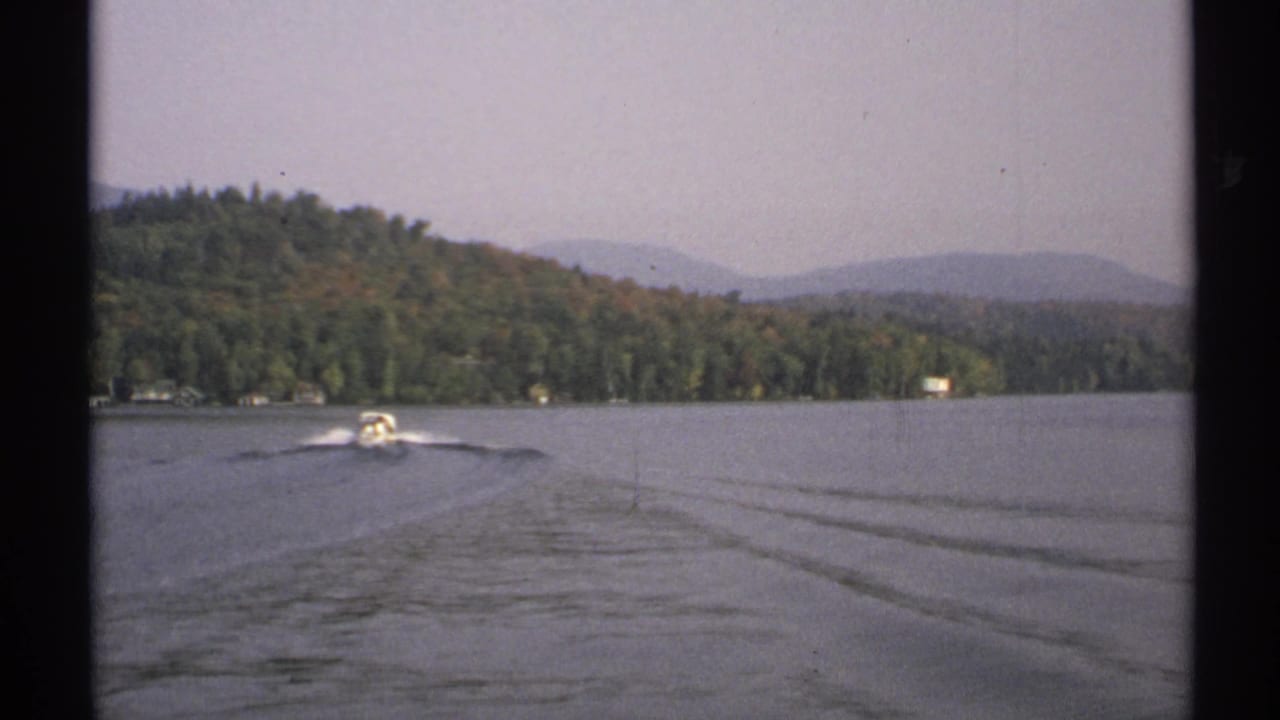 A small motorboat crosses a New York lake in 1971, trailing a bright wake below forested hills and a hazy sky.