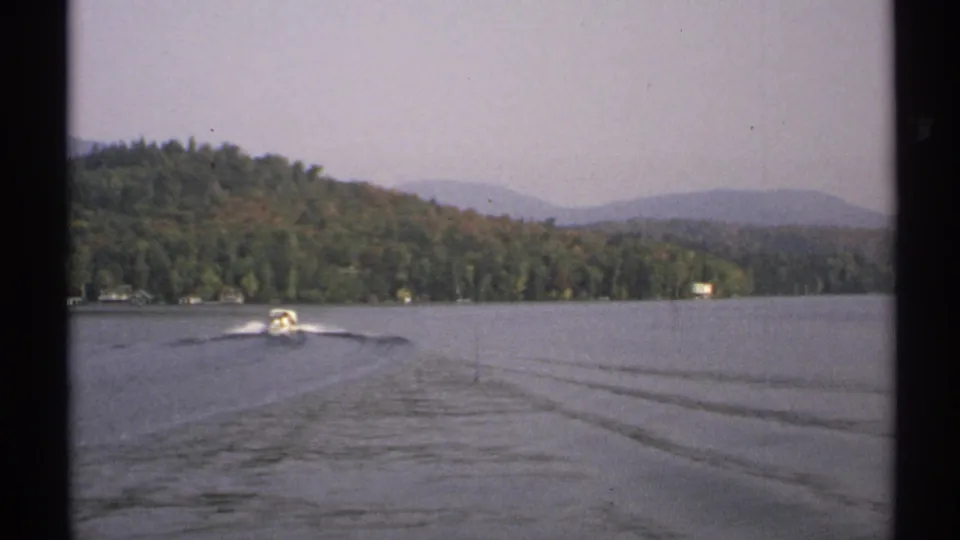A small motorboat crosses a New York lake in 1971, trailing a bright wake below forested hills and a hazy sky.