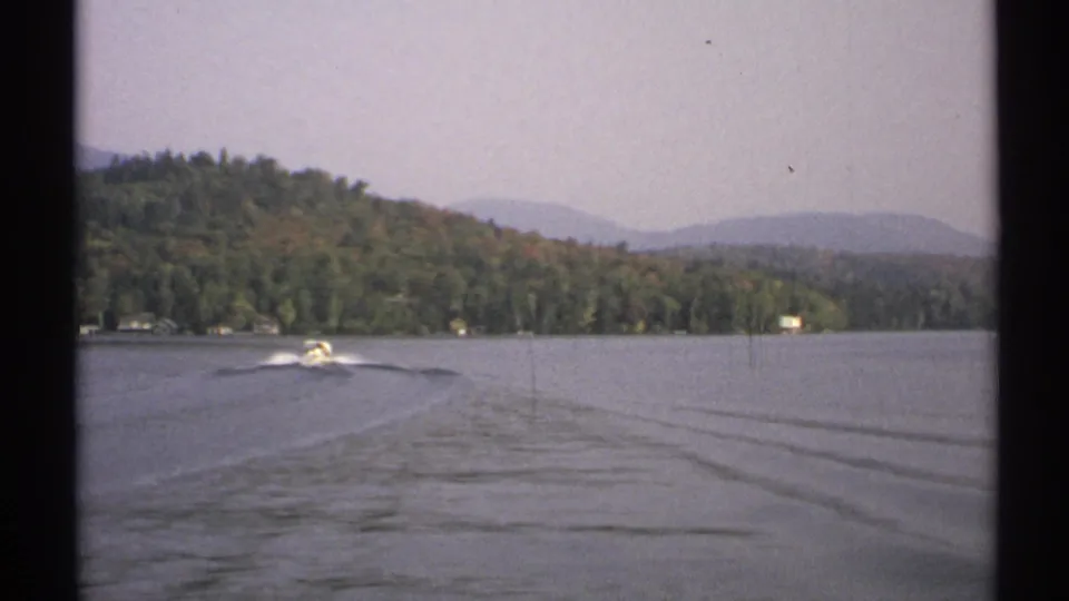 The 1971 8mm footage shows a motorboat pulling away across the lake, with wake lines leading back toward a wooded New York shoreline.
