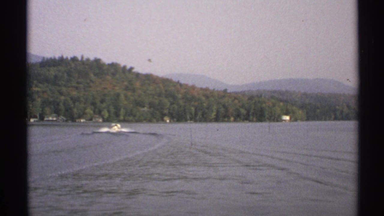 A small motorboat continues across a 1971 New York lake, framed by open water, long wake lines, forested hills, and hazy mountains.