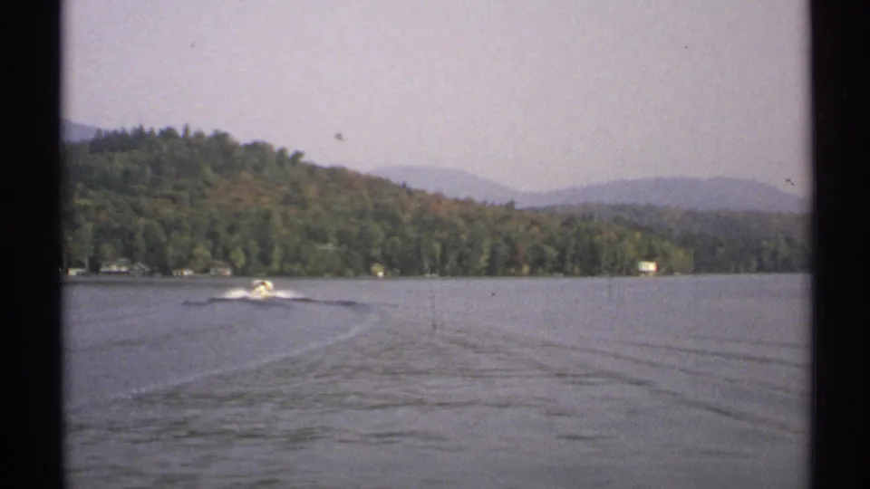 A small motorboat continues across a 1971 New York lake, framed by open water, long wake lines, forested hills, and hazy mountains.