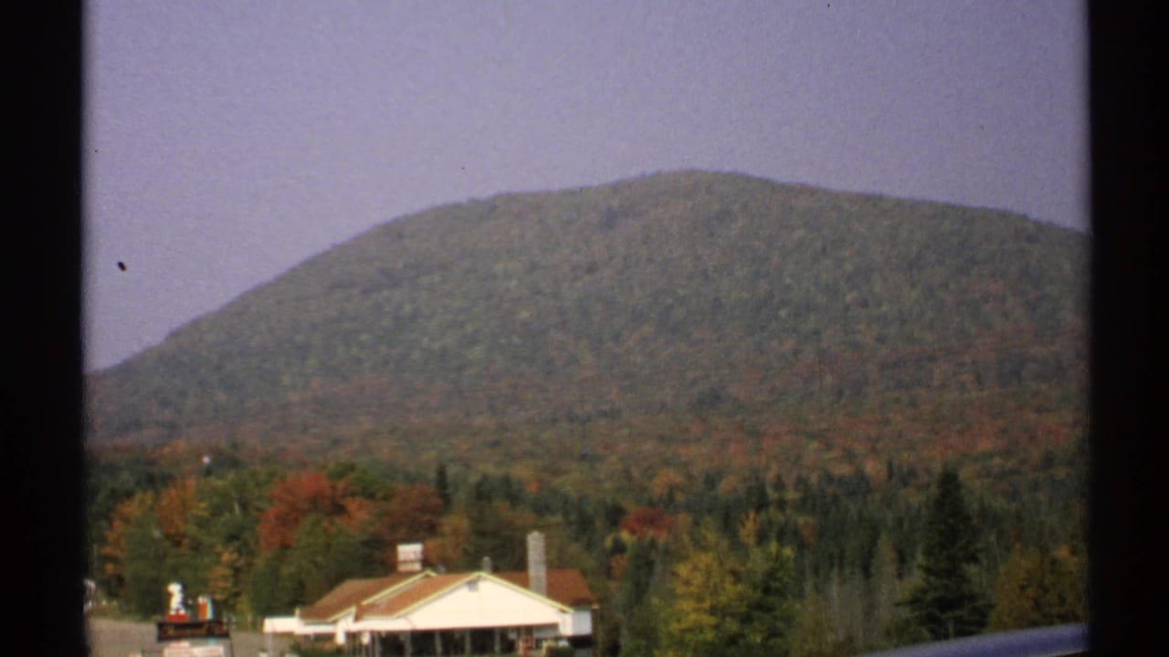 1971 New York 8mm frame showing a forested mountain, autumn treetops, pale sky, and a small building in the foreground.