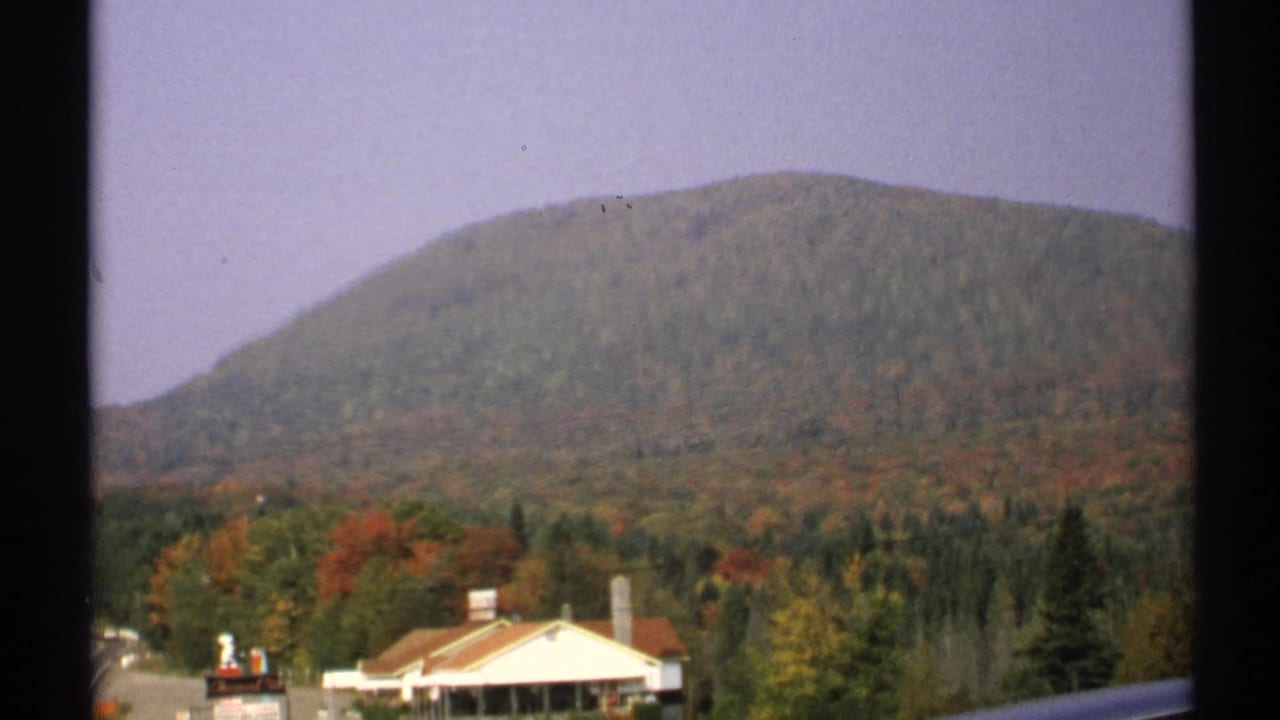 1971 New York 8mm frame showing a forested mountain, early autumn foliage, pale sky, and a low building near the base.