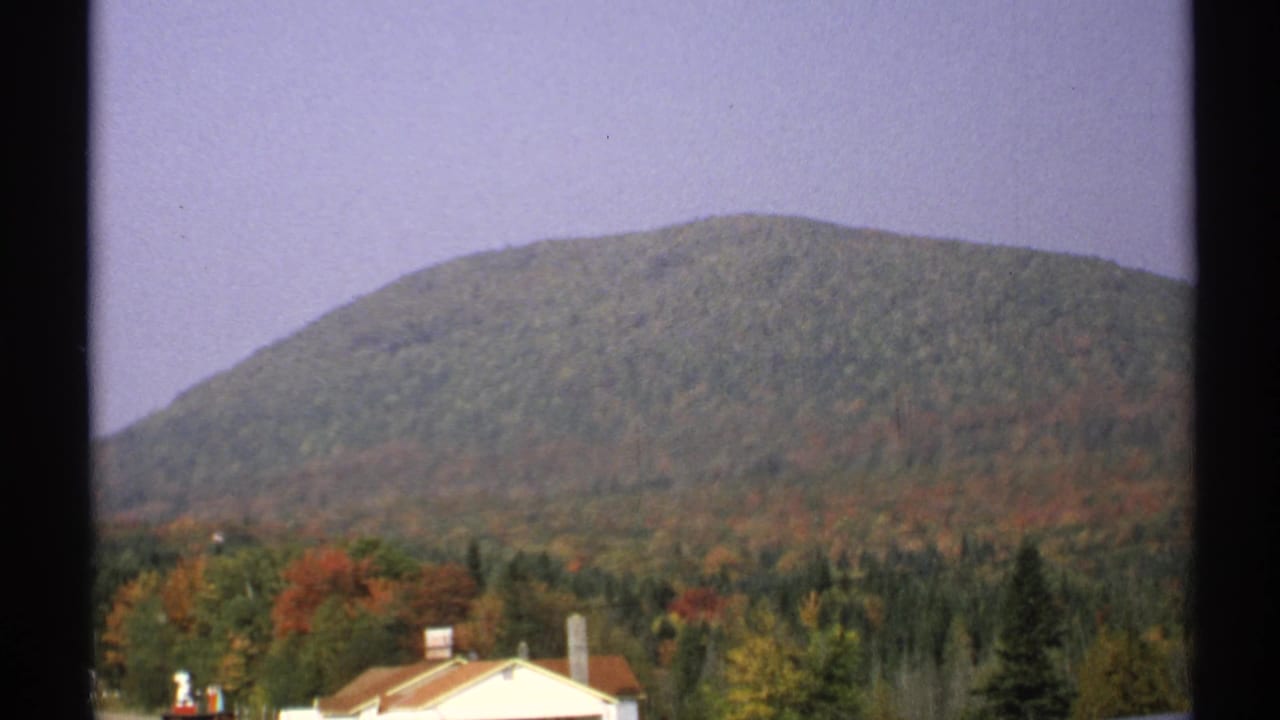 1971 New York 8mm view of a rounded forested mountain above fall-colored trees and a partly cropped building.