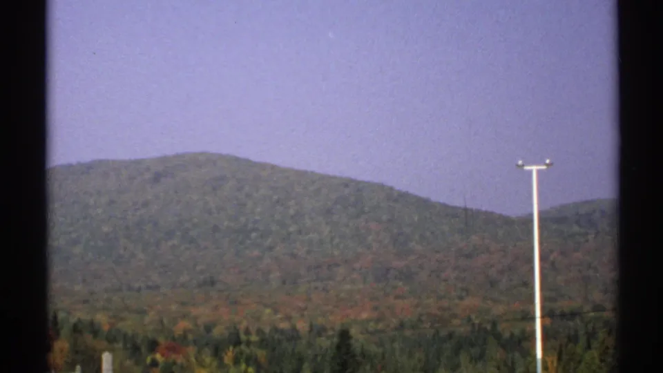 1971 New York 8mm frame with distant wooded hills, a band of autumn foliage, pale sky, and a tall utility pole in the foreground.