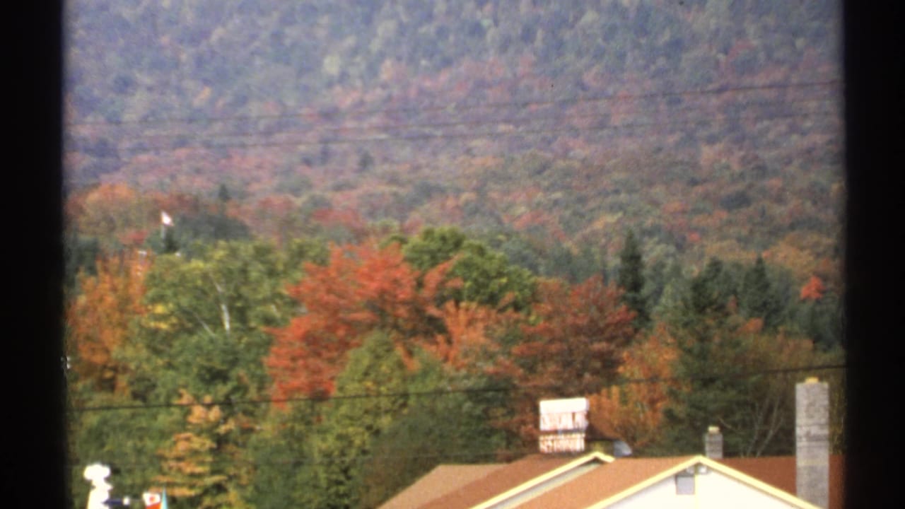 1971 New York 8mm frame with residential rooftops and chimneys in front of fall trees and wooded hills.