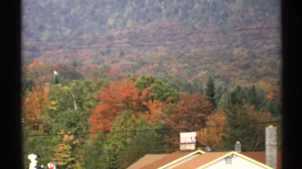 1971 New York 8mm frame with residential rooftops and chimneys in front of fall trees and wooded hills.