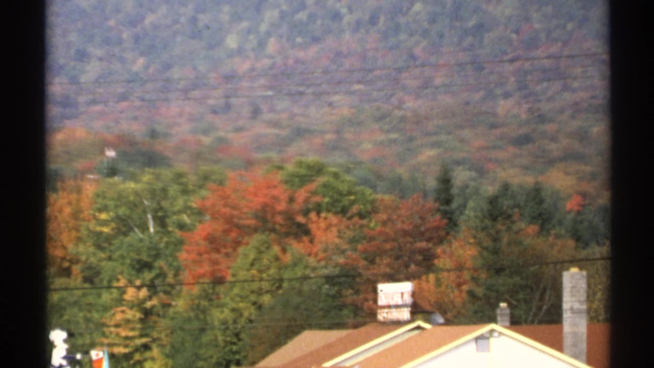 1971 New York home-movie view of fall-colored hills, trees, rooftops, chimney, and utility lines through a window frame.