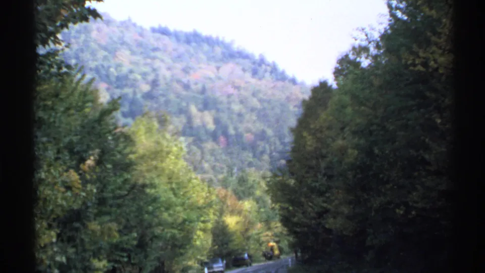 1971 8mm footage frames a moving-vehicle view of roadside trees, a distant road, and a forested New York mountain slope.