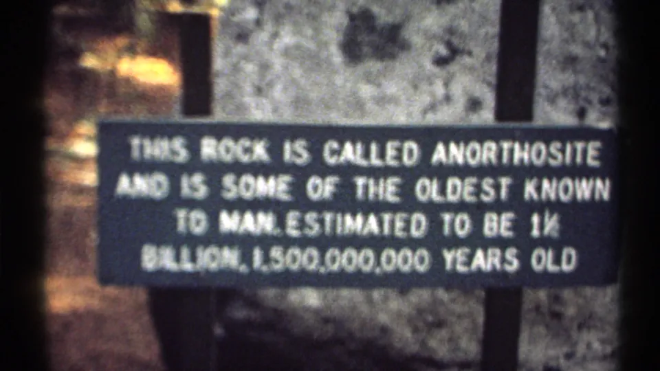 Close 8mm view of the anorthosite sign, with white text, a gray rock backdrop, and muted wooded surroundings.