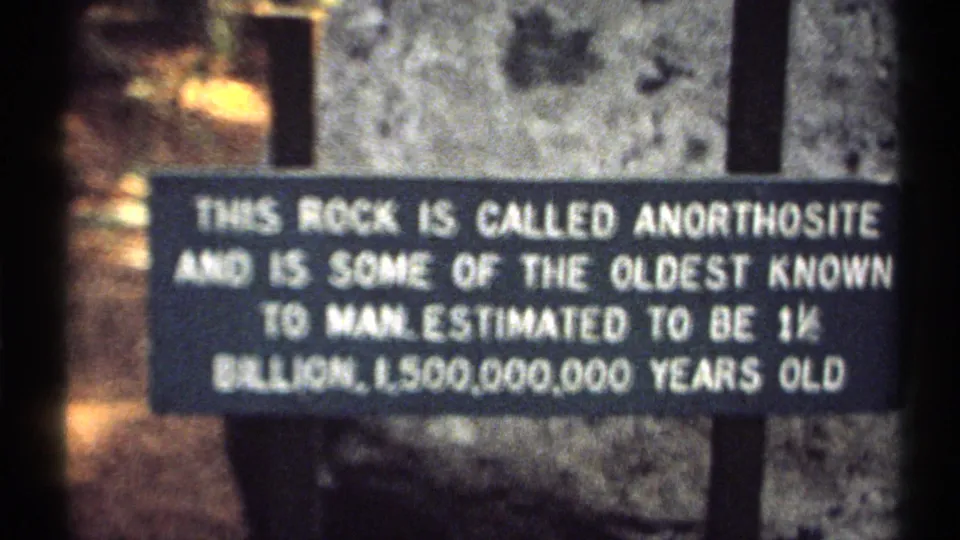 Home-movie frame showing the anorthosite display sign in soft focus, with aged film color and a rocky background.