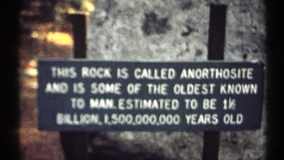 Final 8mm frame of the black anorthosite sign, gray rock, and dark supports in a wooded outdoor setting.
