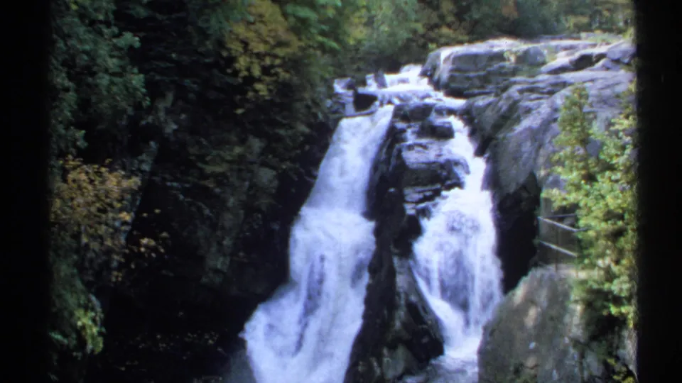 1971 New York 8mm frame showing white water cascading over dark rock ledges with dense greenery around the falls.