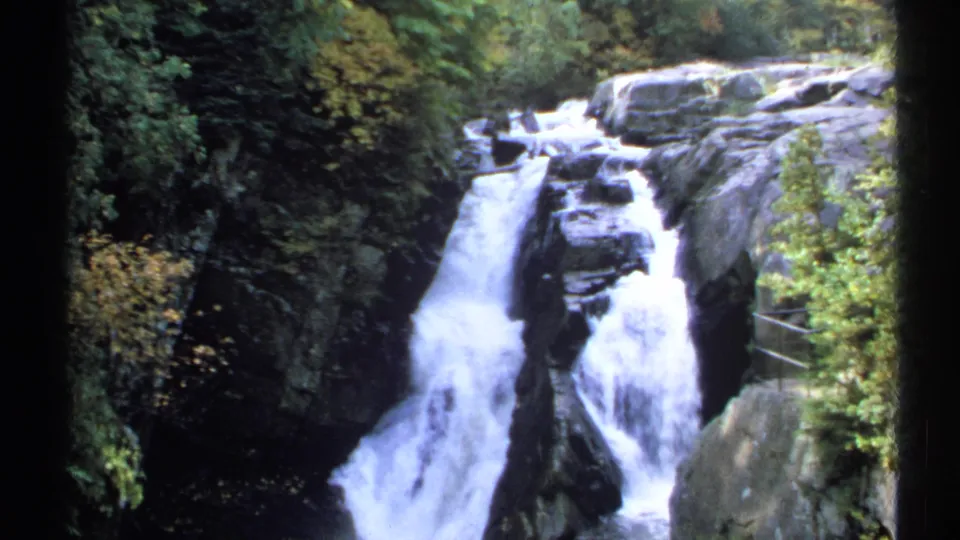 1971 New York home-movie frame of a waterfall spilling over dark stone, bordered by trees, shrubs, and a small railing-like structure.
