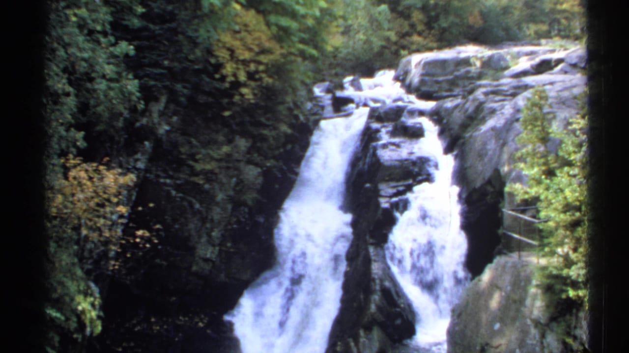 1971 New York 8mm frame of white water dropping between dark rocks, with dense greenery and a railing at the right.