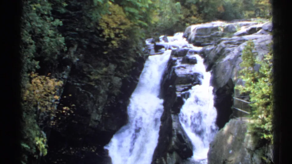 1971 New York 8mm frame of white water dropping between dark rocks, with dense greenery and a railing at the right.