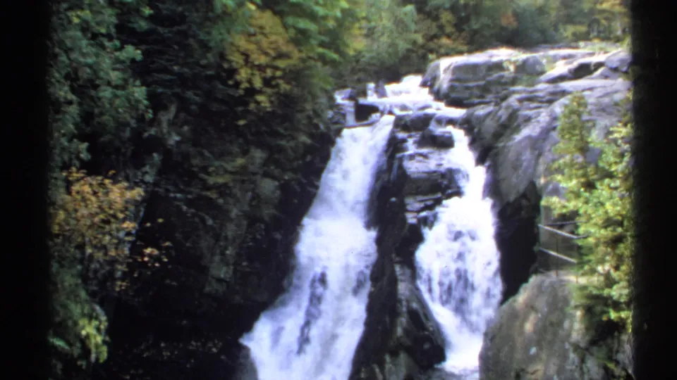 1971 New York 8mm frame showing the waterfall from a lookout angle, with mossy rocks, foliage, and a metal rail.