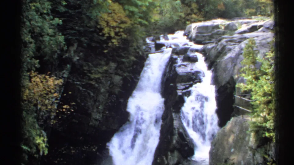 1971 New York 8mm frame of cascading water split by a central rock, surrounded by trees and a right-side viewing rail.