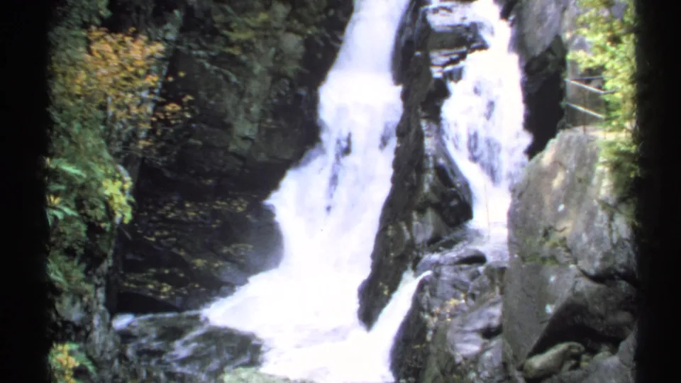 1971 New York 8mm frame with a closer view of the falls, dark wet rocks, foaming water, and a partial rail at right.