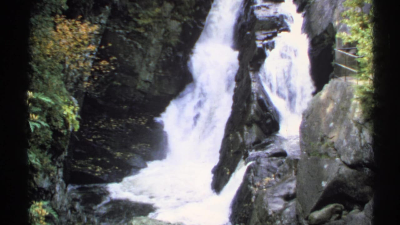 1971 New York 8mm frame of white water falling through dark rocks, bordered by green foliage and yellow leaves.