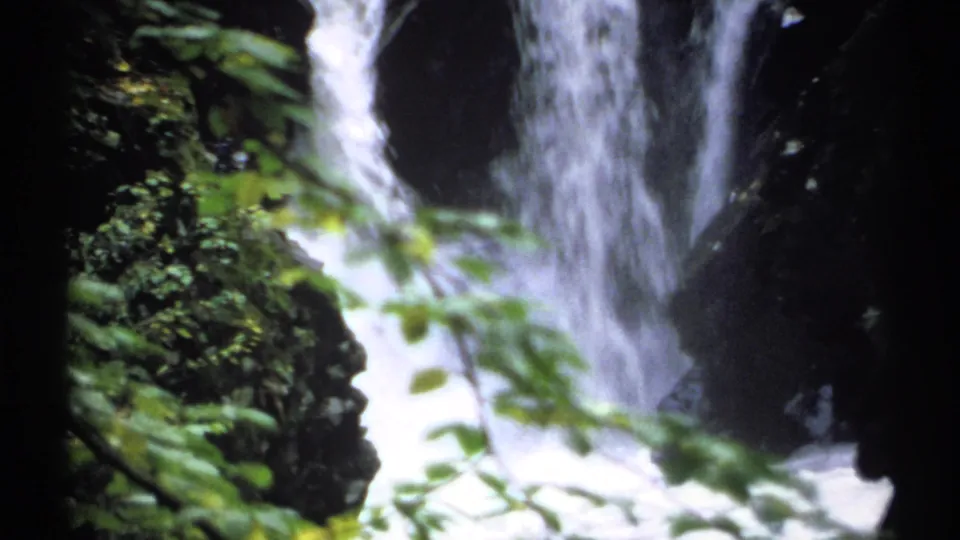 1971 New York 8mm frame showing foaming waterfall flow in a narrow rocky canyon, partially obscured by green leaves.