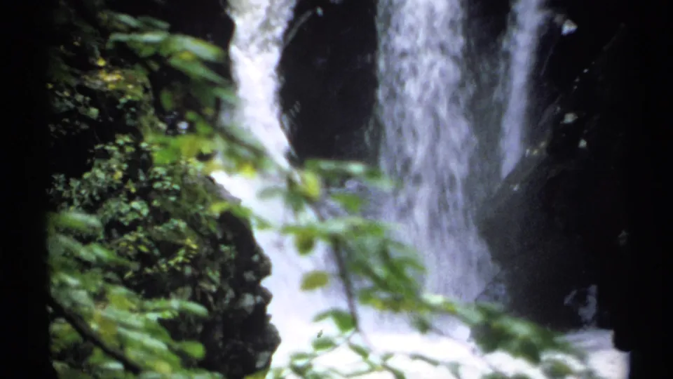1971 New York 8mm frame of a waterfall cascading through a shaded forest gorge, with leaves blurred across the foreground.