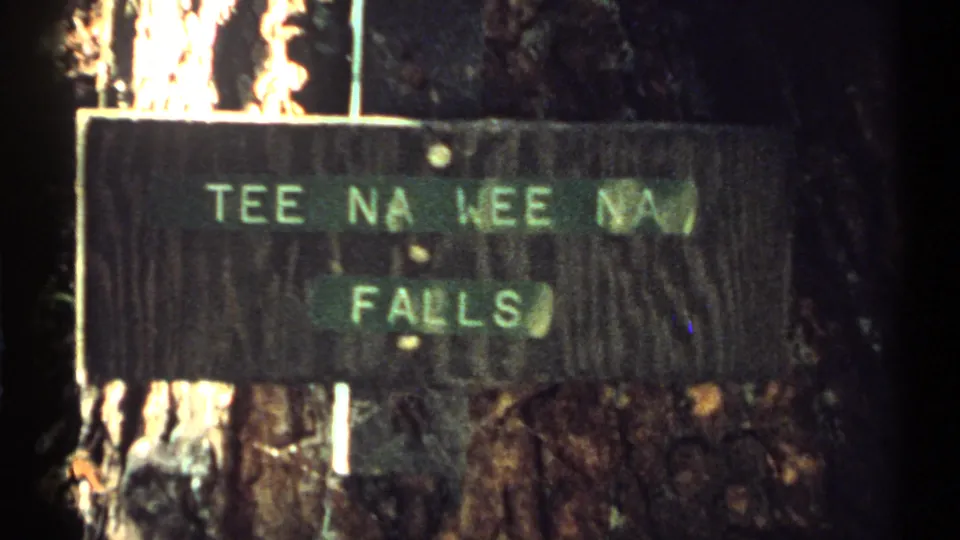 Close 1971 New York 8mm view of the Tee Na Wee Na Falls sign, with painted lettering on weathered wood and tree bark behind it.
