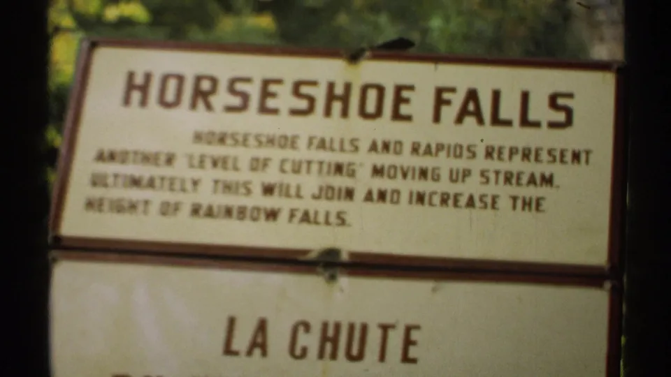 1971 New York 8mm frame showing a Horseshoe Falls interpretive sign with a lower bilingual panel and green foliage in the background.