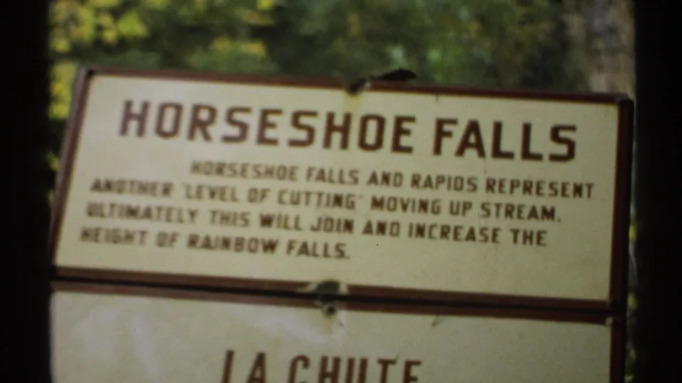 1971 New York 8mm frame showing a slightly off-center Horseshoe Falls sign above a La Chute panel, with foliage behind the boards.