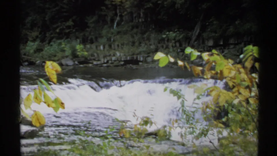 1971 New York 8mm frame showing bright white water, foreground leaves, rocks, and a shadowed forest backdrop.