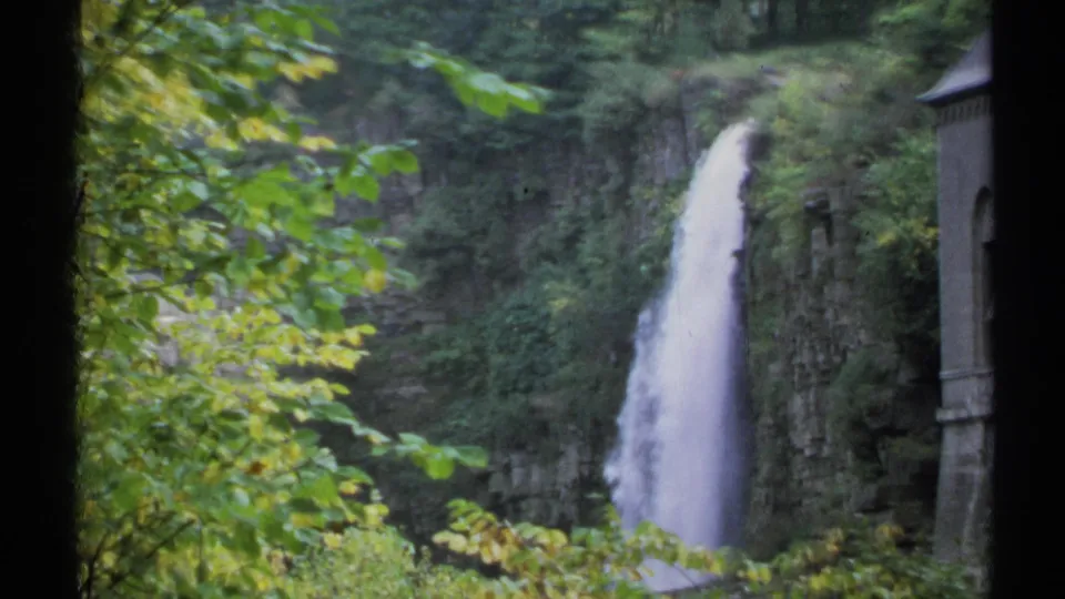 1971 New York 8mm frame showing a tall waterfall, rocky gorge, green foliage, and part of a stone structure at right.