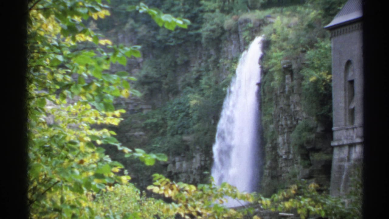 Waterfall and cliff face seen through foliage, with a stone building edge at right.