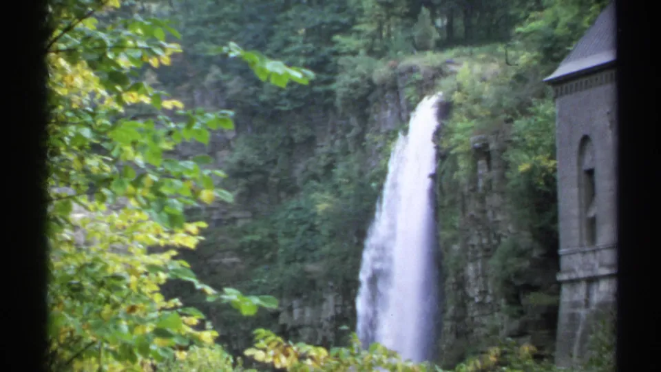 1971 New York 8mm frame showing the waterfall from a visitor viewpoint, with foreground leaves, cliff rock, and stonework.