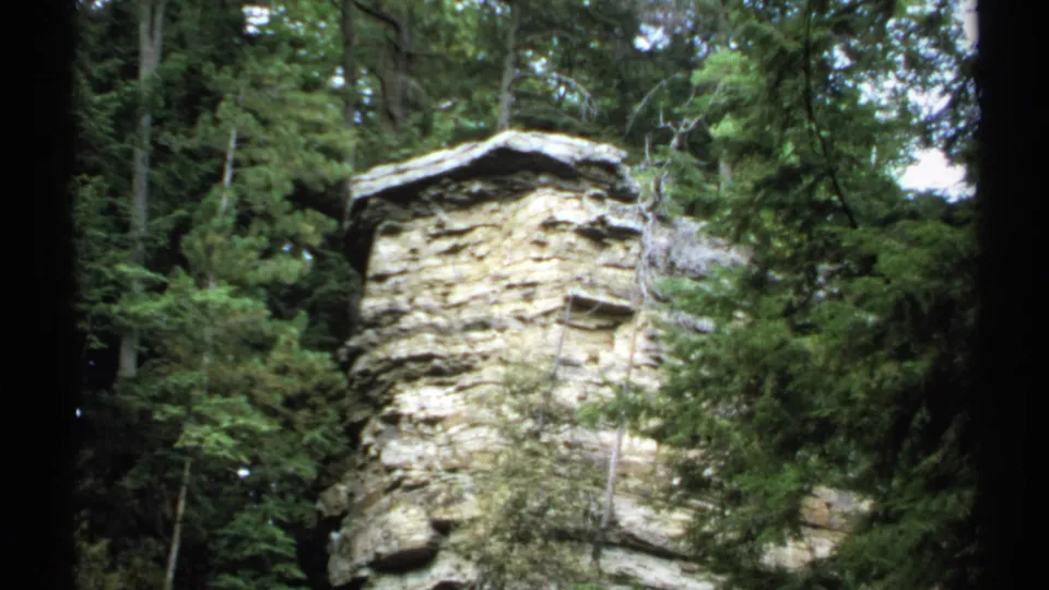 1971 New York 8mm frame showing a layered rock outcrop surrounded by trees and dense green vegetation.