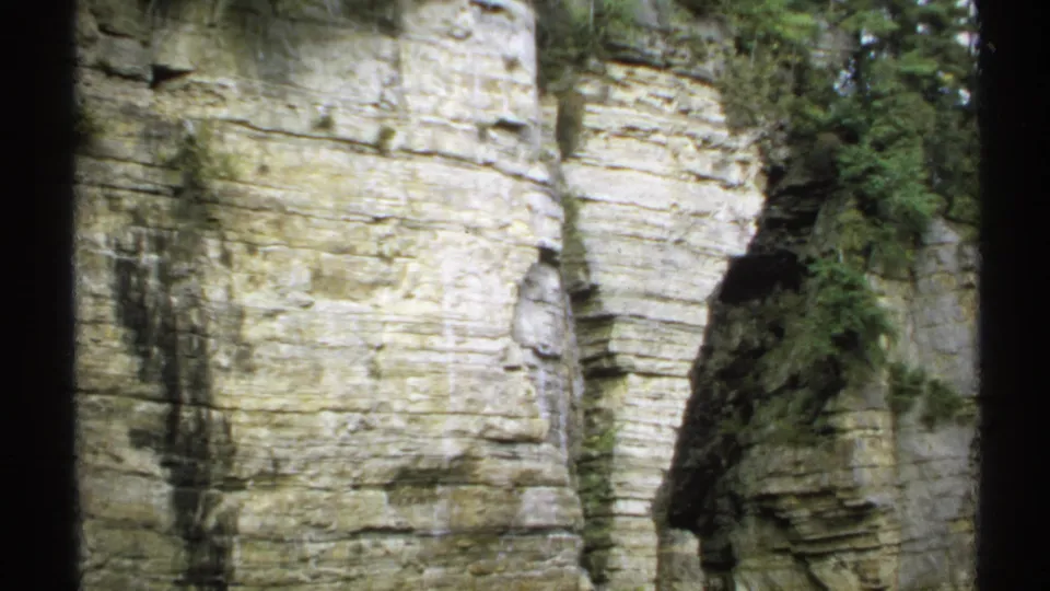 Closer 1971 8mm view of a layered pale rock wall with forest trees along the edge of the frame.