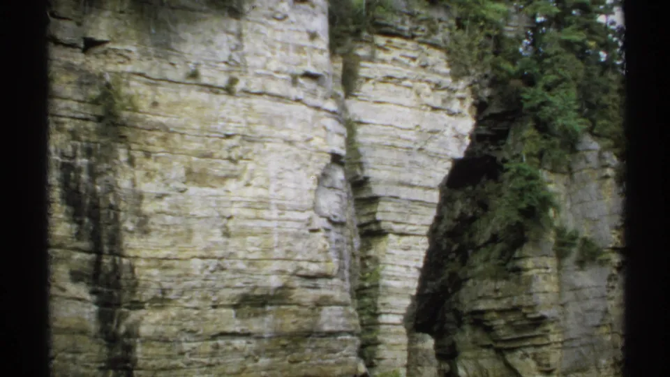 1971 home-movie frame showing a pale layered cliff face in New York, with a narrow vegetated seam and soft film grain.