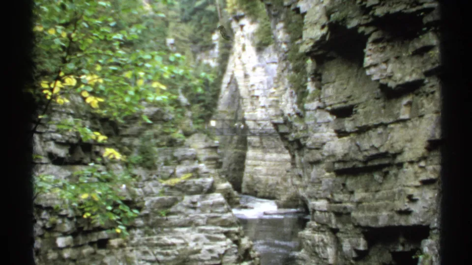 1971 New York 8mm frame showing layered rock walls, green foliage, a narrow stream, and a small pedestrian bridge in the distance.