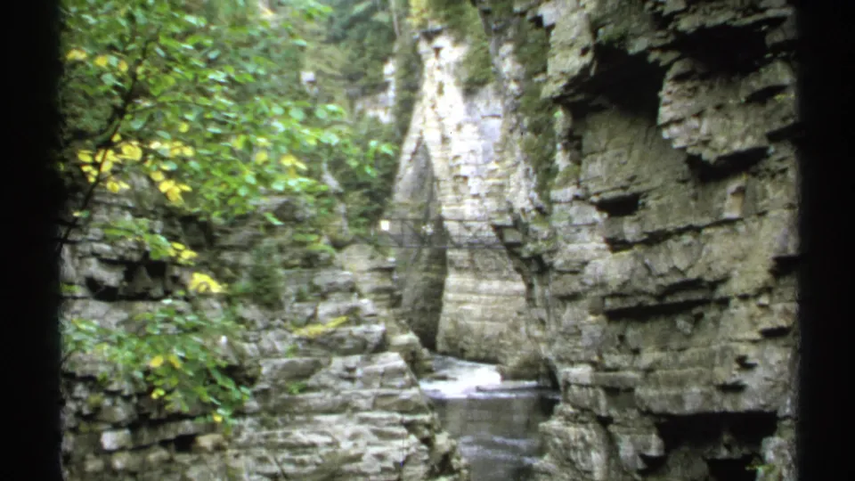 1971 New York home-movie view of a rugged gorge, with stratified rock, leafy trees, moving water, and a pedestrian bridge set back in the scene.