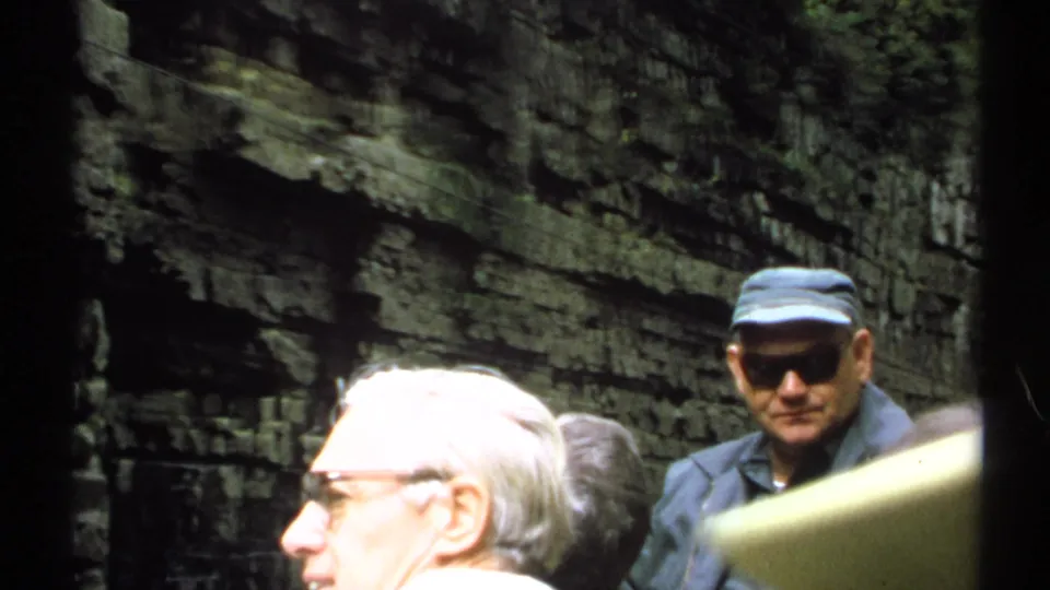 Final 1971 New York frame showing two adult men at a gorge overlook, one in a cap and sunglasses, with layered rock cliffs behind them.